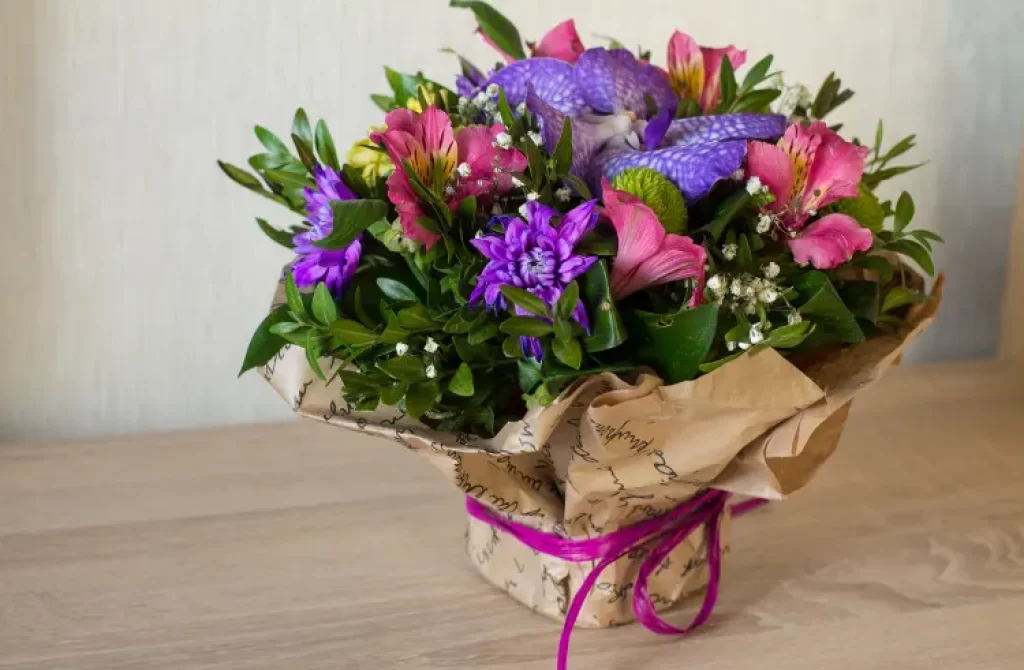 Vibrant mixed flower bouquet wrapped in patterned paper, secured with a purple ribbon, on a wooden surface.