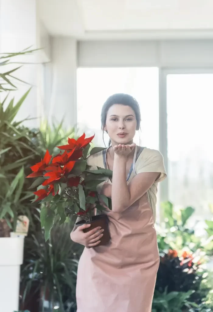 A woman holding a potted plant with bright red flowers in an indoor setting, surrounded by other plants.